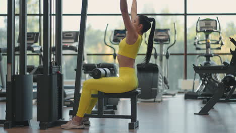 Hispanic-woman-sitting-on-a-simulator-in-the-gym-pulls-a-metal-rope-with-the-weight-pumps-up-the-muscles-of-the-back.-brunette-woman-pulls-on-simulator.-performing-exercise-for-back-muscles-simulator